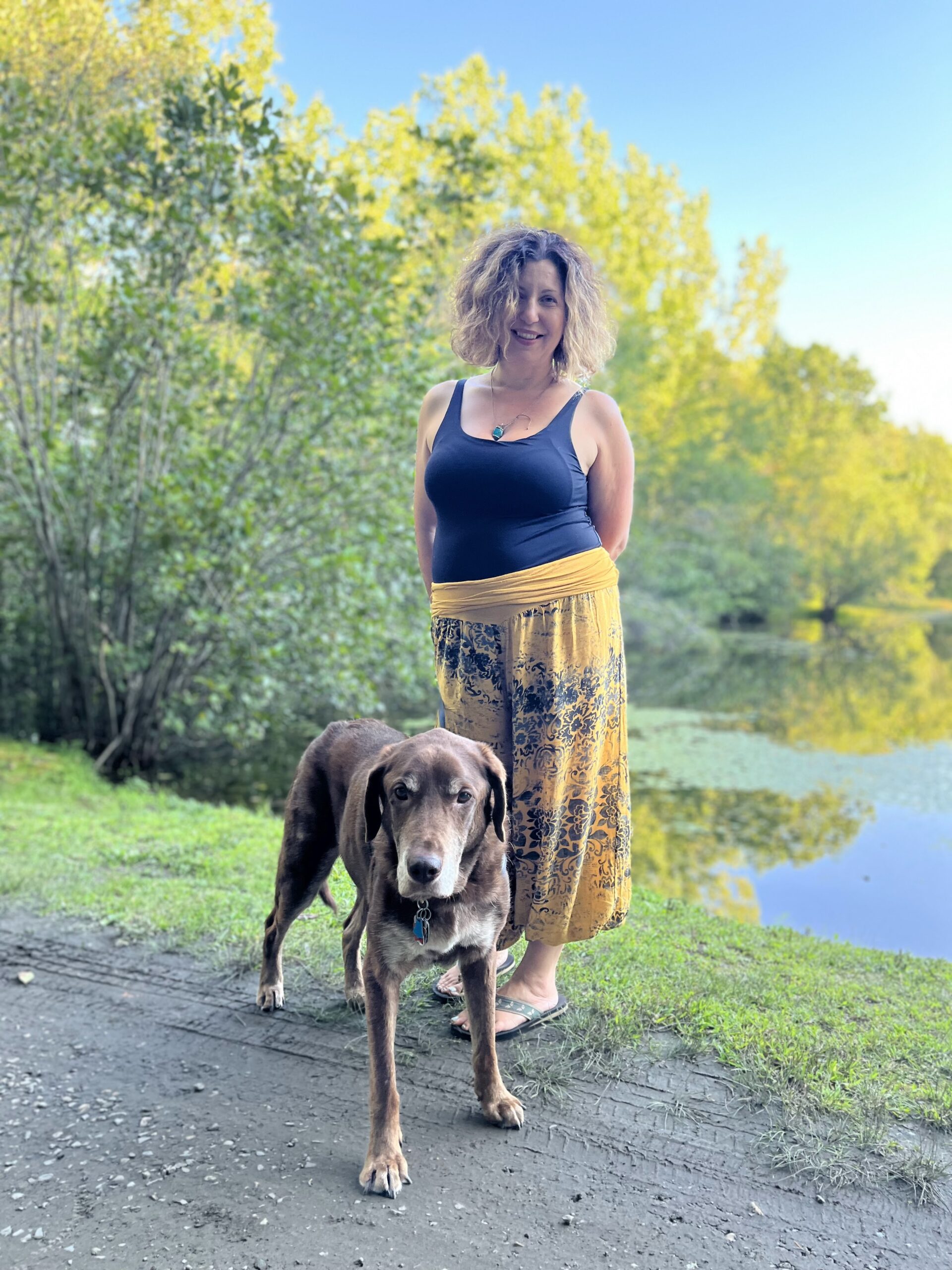 Jen walking Reed in the woods Woman in navy blue tank top with yellow flowery pants on a walking path by a lake in the woods with her white and brown dog.