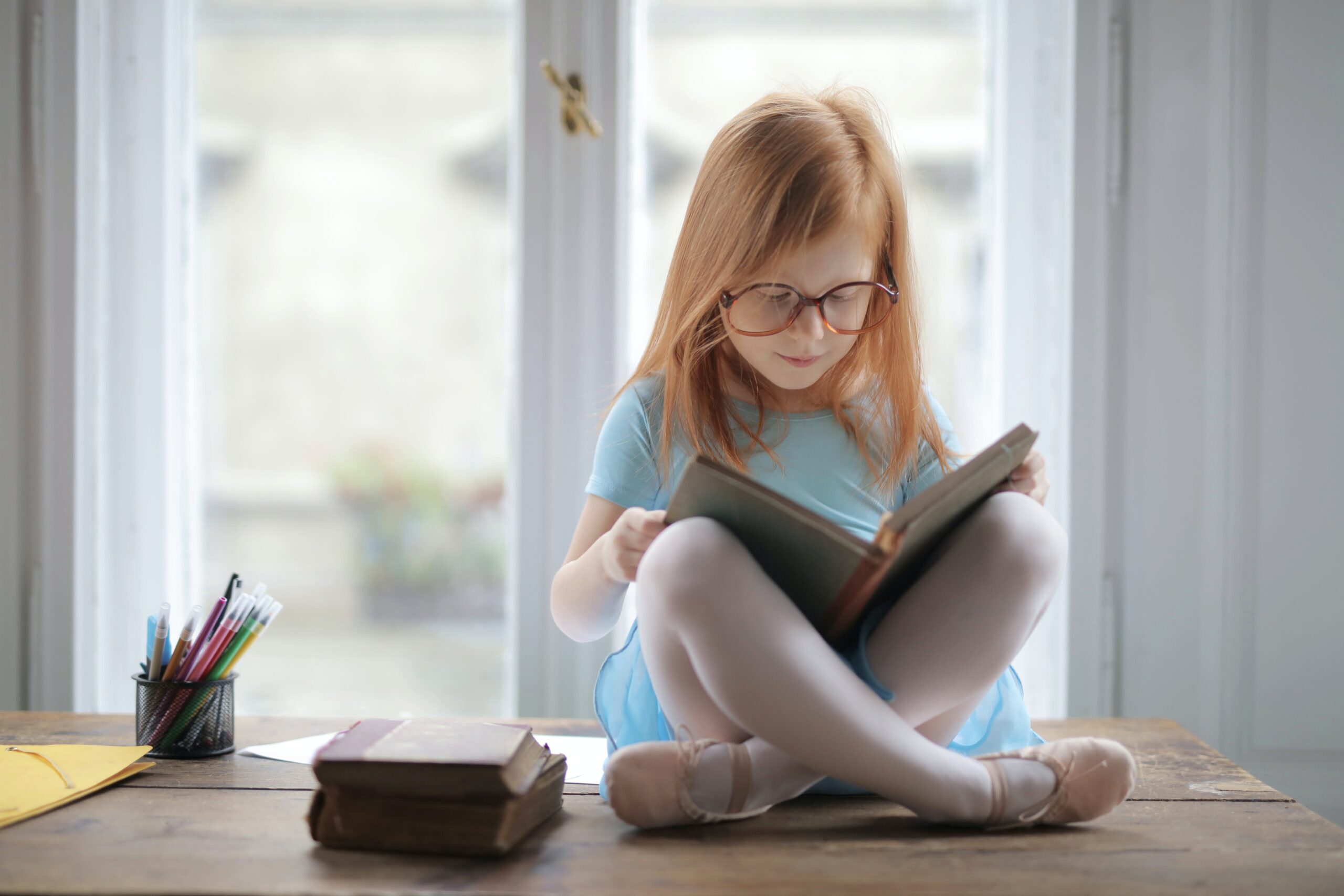 Young girl with long strawberry blond hair wearing glasses reading a book on her lap as she sits criss-cross applesauce on a table with 2 closed books and a cylinder pencil holder in front of a window.